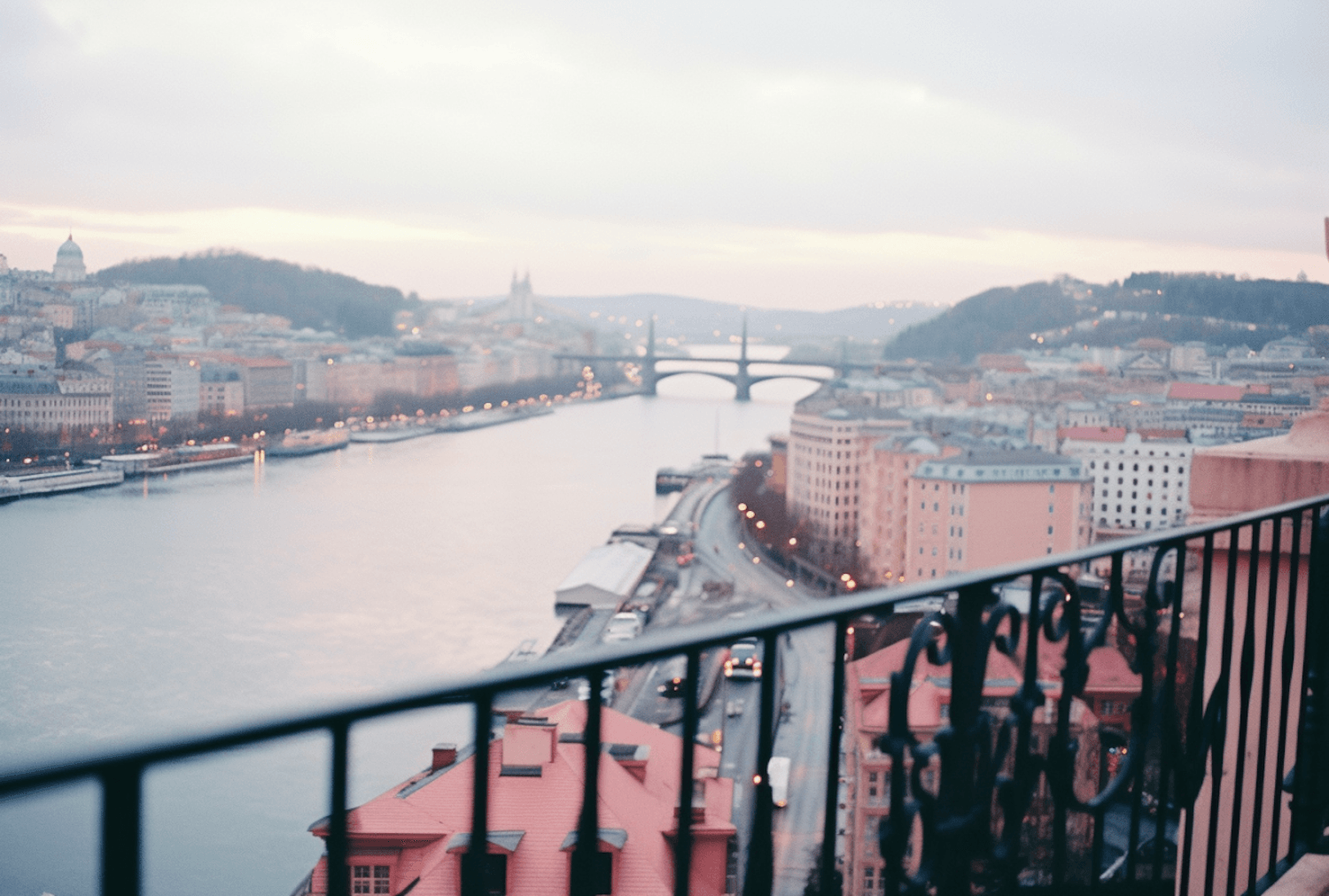 A wide river winding through a European city at dusk, its bridge and hillsides lit up