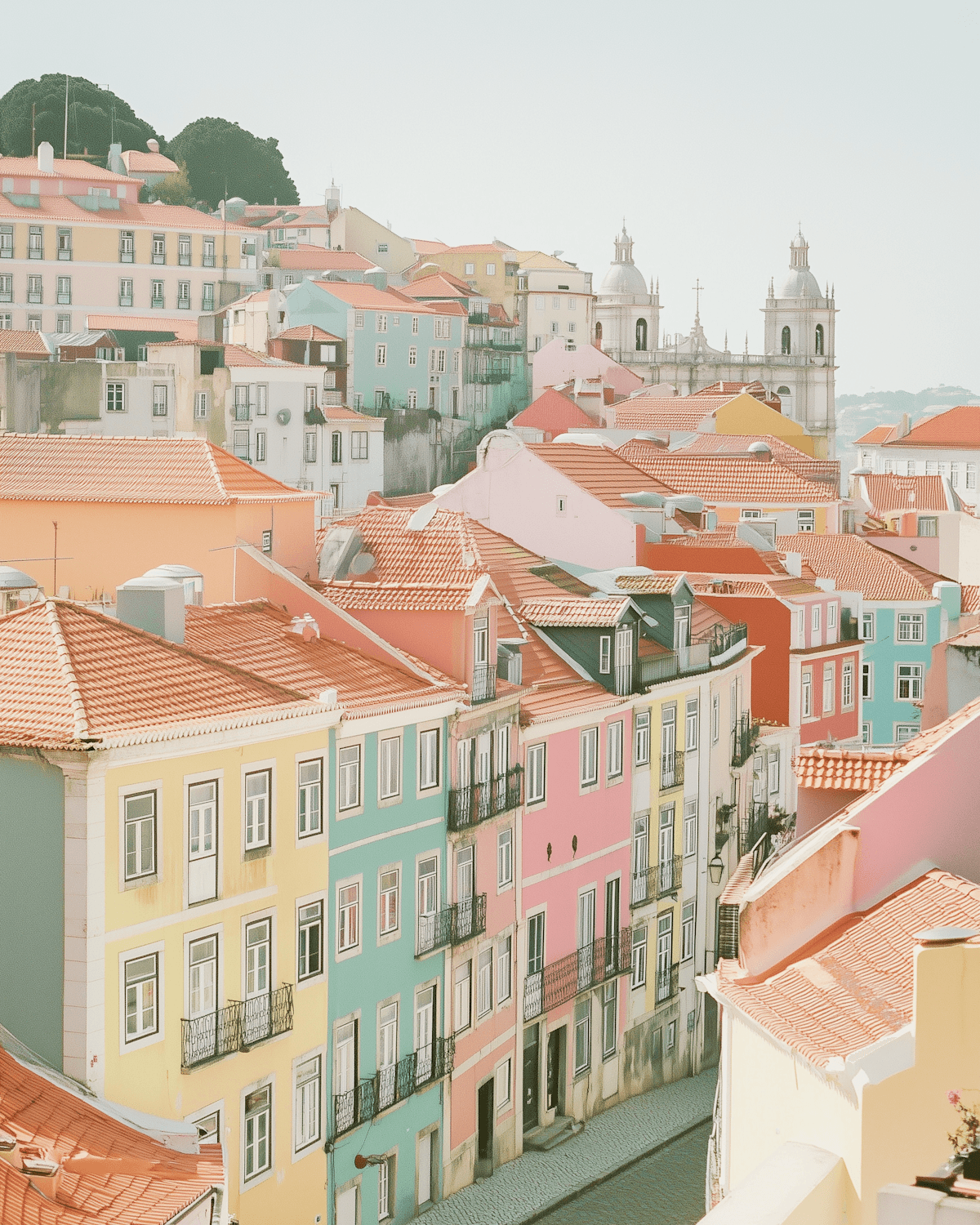 Pastel houses and terracotta rooftops climbing a hill in Lisbon, with a white church on the skyline